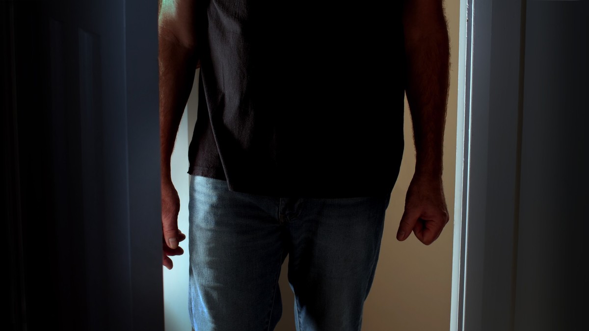 Silhouette of a man opening a door to a interior dark room, possibly a bedroom. He is an unrecognisable figure standing in a doorway clenching his fist.