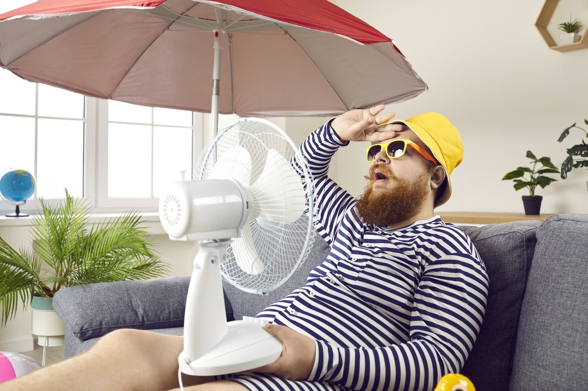 Funny sweaty chubby bearded man in swimsuit sitting at home, suffering from crazy summer heat, wiping sweat off forehead, holding electric fan, wishing for heatwave to stop and fresh breeze to blow