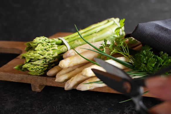 Fresh green and white asparagus on wooden cutting board against black stone wall. Vegetable for healthy nutrion concept. Lifestyle cooking background. Close-up with hort depth of field and space for t ...