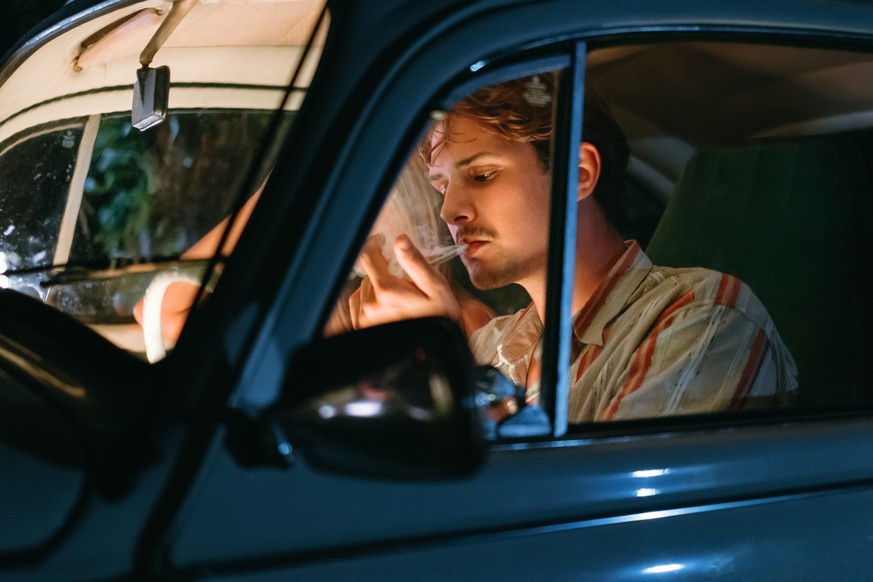 Young man smoking marijuana joint while sitting beside woman in vintage car at night