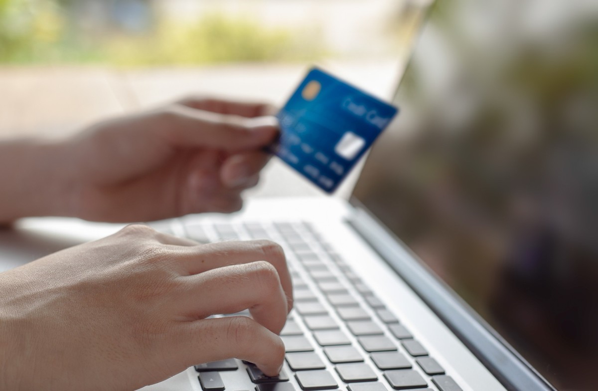 Woman hand holding blue credit card and using a modern laptop, typing and texting. against defocus green nature outdoor. mobile payment, online shopping concept