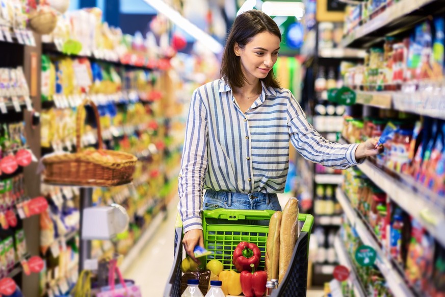 At The Supermarket. Portrait Of Young Woman Standing With Trolley Cart Between Aisles In Grocery Store. Cheerful Consumer Buying Essentials In Local Shop, Taking Products From Shelf