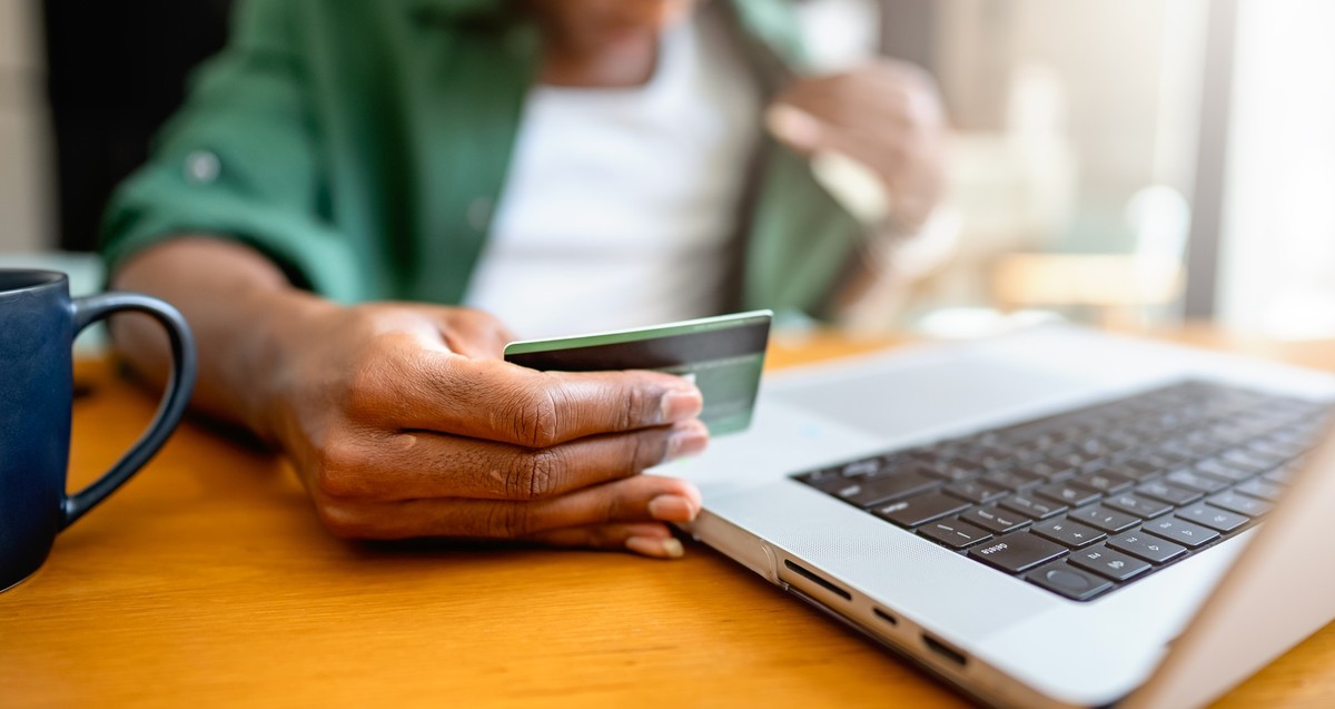 A young man of African descent sits by a wooden table in a modern, sunlit living room, smiling as he uses a laptop to make an online purchase and pays by credit card.
