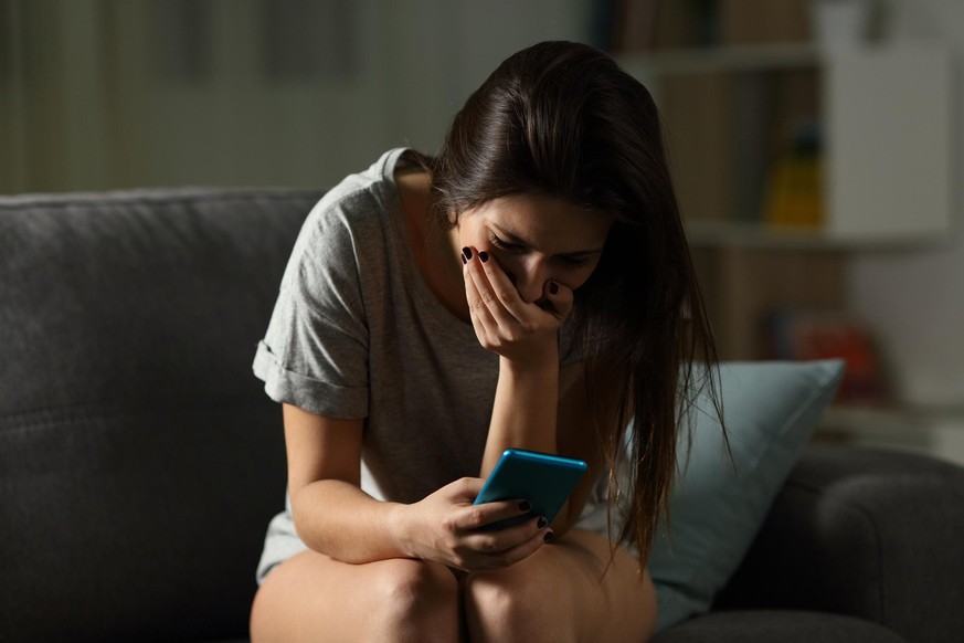 Sad teen receiving bad news online in a smart phone sitting on a couch in the living room at home