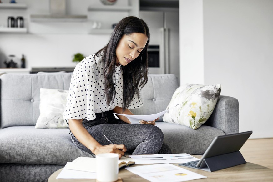 Young woman analyzing bills while writing in diary. Beautiful female is using digital tablet at table. She is sitting on sofa at home.