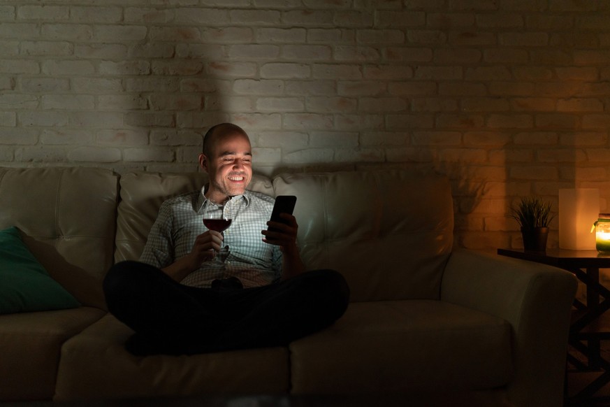 Portrait of a man in his 30s relaxing at home with some wine and checking social media on a smartphone at night