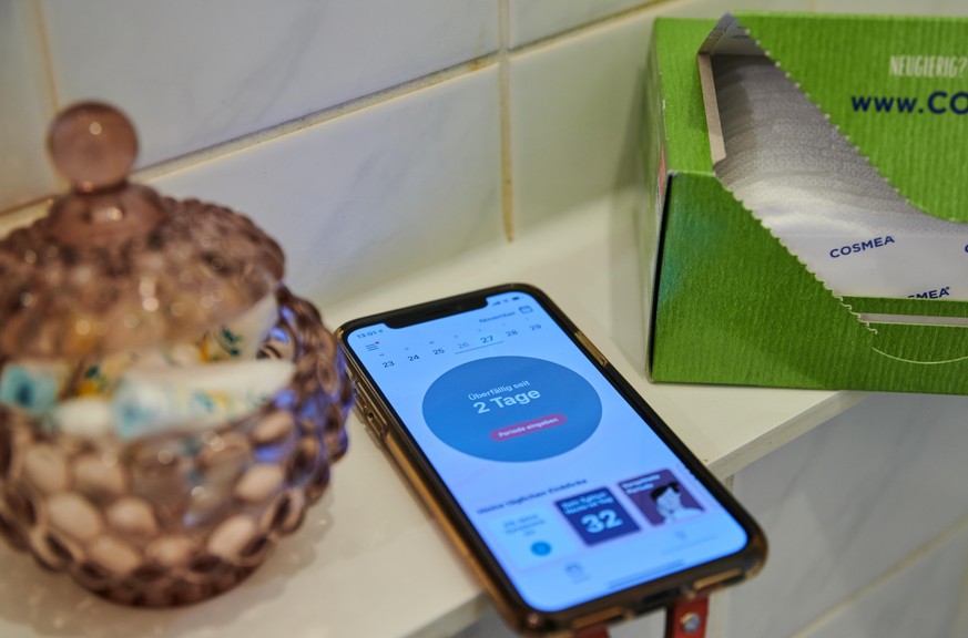 Tampons, Slipeinlagen und ein Smartphone mit einer Menstruations-App liegen im Badezimmer.