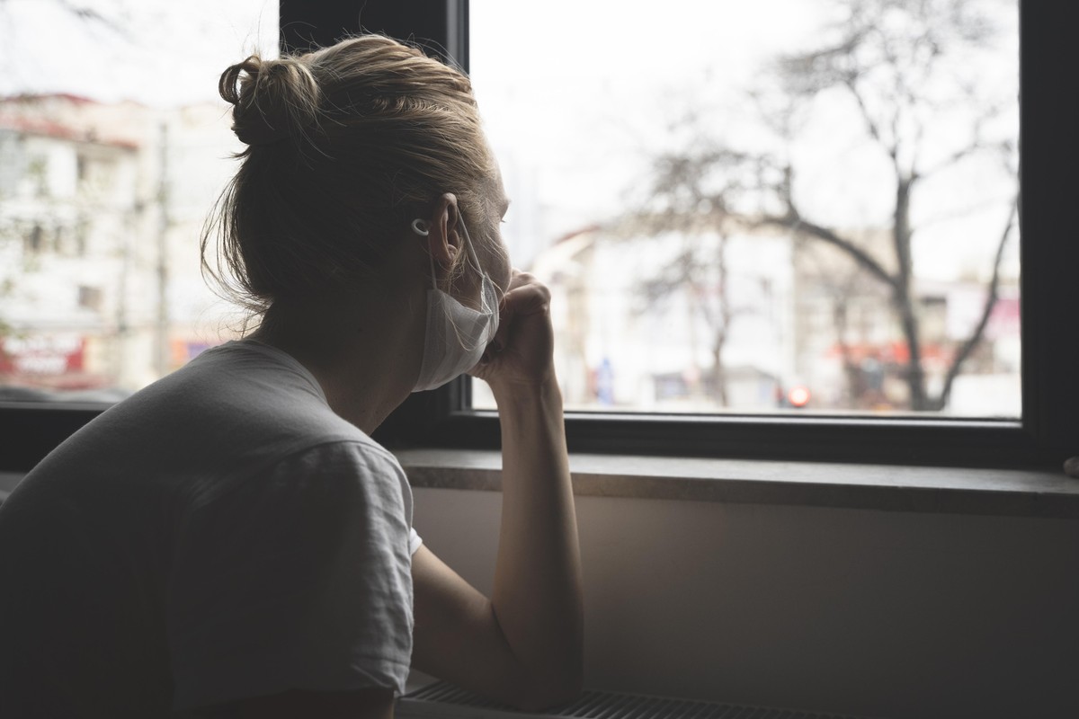 rear view closeup of young Caucasian woman wearing medical protection face mask watching the city by the window quarantine concept