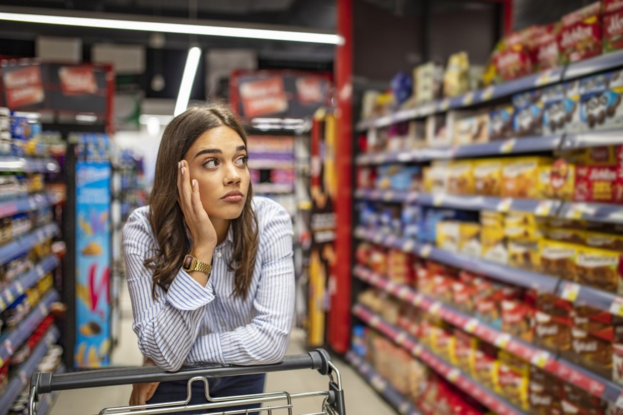 Young woman staying confused in grocery store. Confused woman doesn&#039;t know what to buy. Young woman with a shopping cart at supermarket. Looking at supermarket shelf