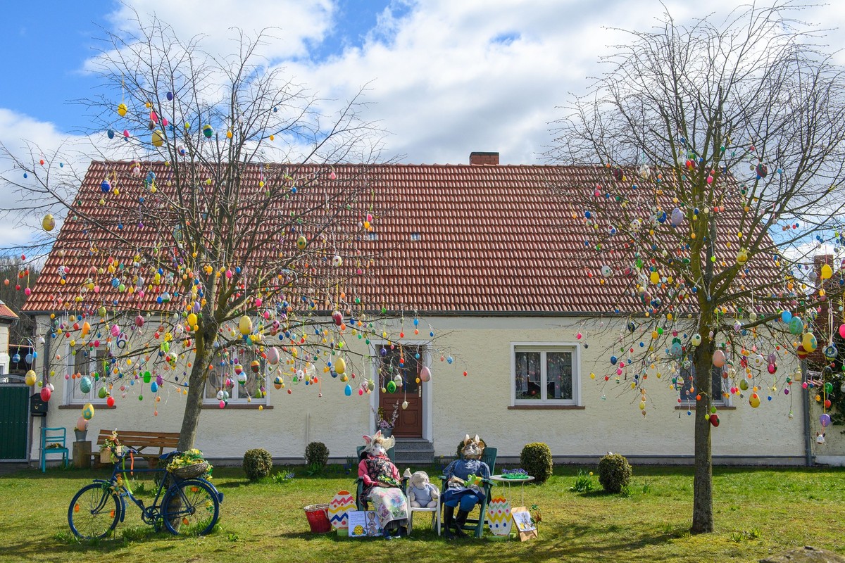 03.04.2023, Sachsen-Anhalt, Wulkau: Ostereier h�ngen vor einem Haus in den �sten von zwei Linden. Darunter sitzen Puppen, die ein Osterhasenpaar darstellen. Die bunten Eier waren etwa f�nf Wochen vor  ...