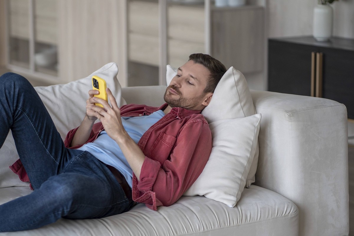 Desinteressierter Mann mit Telefon auf der Couch liegend surfen online Web, Prokrastination zu Hause. Faul männlich mit Social Media Sucht auf Smartphone scrollen Internet, genießen Sie das virtuelle ...