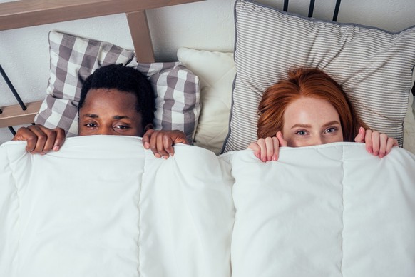 redhaired ginger caucasian happy female and multi-ethnic afro man together lying in bed bedroom hiding under the blanket.bashfulness concept.