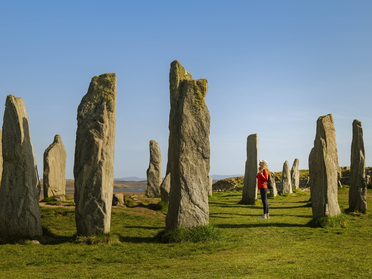 A young tourist admires the ancient Callanish Stones on the Isle of Lewis, Scotland, during a picturesque sunset The historic megaliths stand tall against a clear sky, providing a serene view and a gl ...
