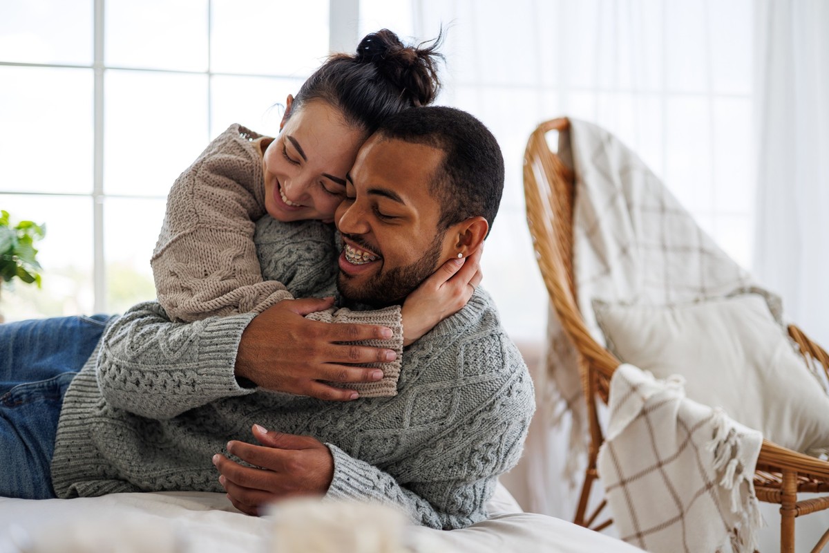 Happy young asian woman in sweater hugging african american boyfriend and relaxing on bed at home	 xkwx african american asian bed bedroom boyfriend cheerful clothing cold weather copy space comfort c ...