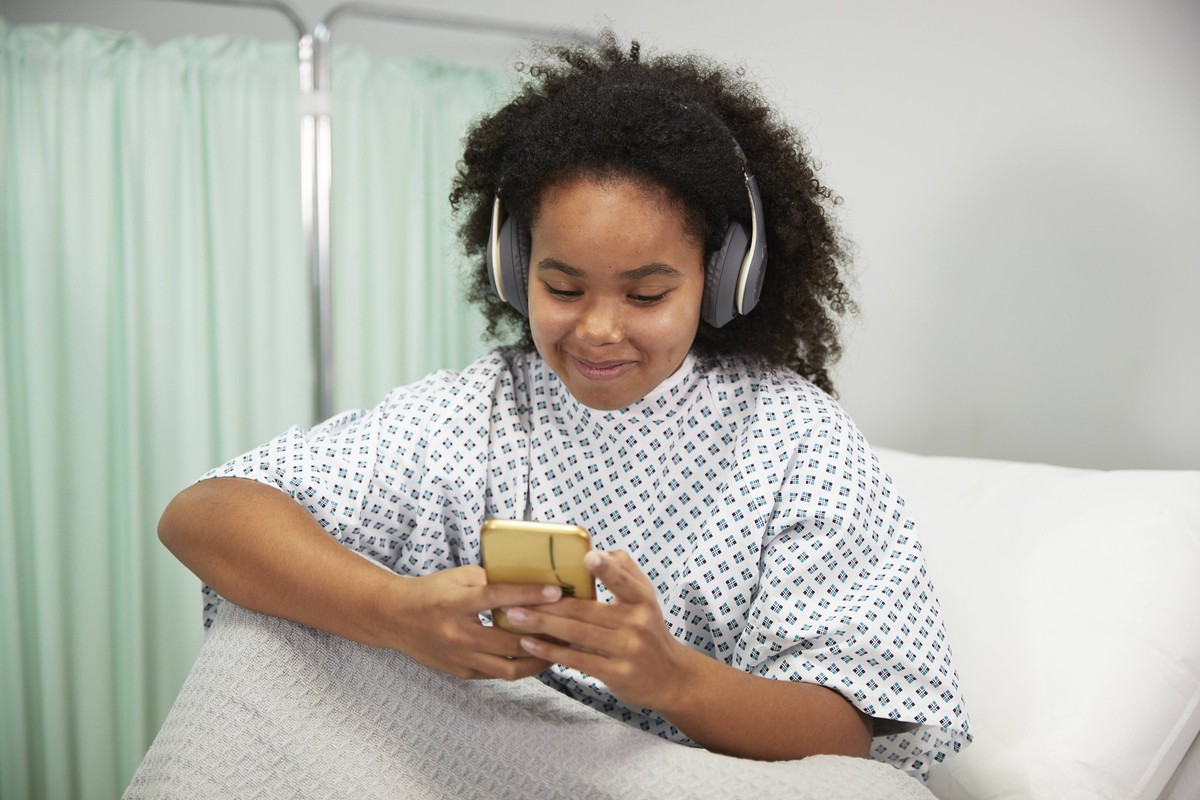 Smiling patient with headphones using smart phone sitting on bed at hospital model released property released, PMF02185