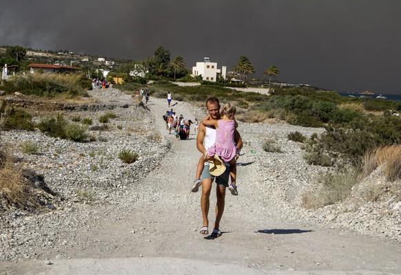 22.07.2023, Griechenland, Rhodos: Ein Mann trägt ein Kind, als sie ein Gebiet verlassen, in dem ein Waldbrand auf der griechischen Insel Rhodos wütet. Ein großes Waldfeuer, das auf der griechischen In ...