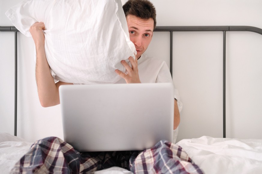 Frightened handsome young man in the white shirt and pajama sitting on a bed with laptop hding behind pillow. Morning of a freelancer