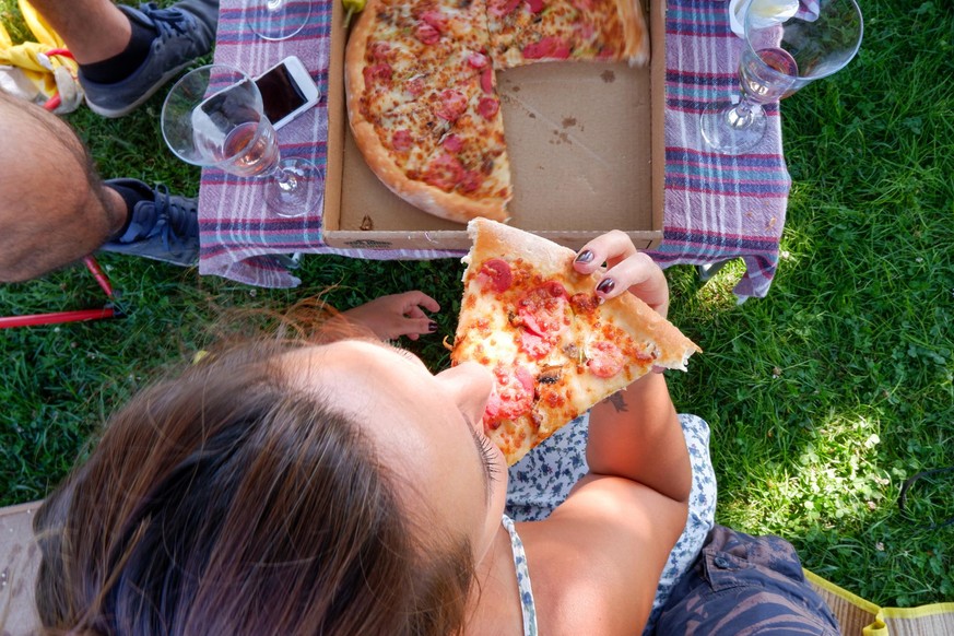 A woman having slice of pizza at picnic with pepperoni pizza and rose wine on city park outdoor with friend in summertime. Flat lay. A pleasant weekend city activity.
