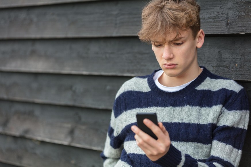Sad depressed thoughtful male boy teenager blond child outside using mobile cell phone for texting or social media