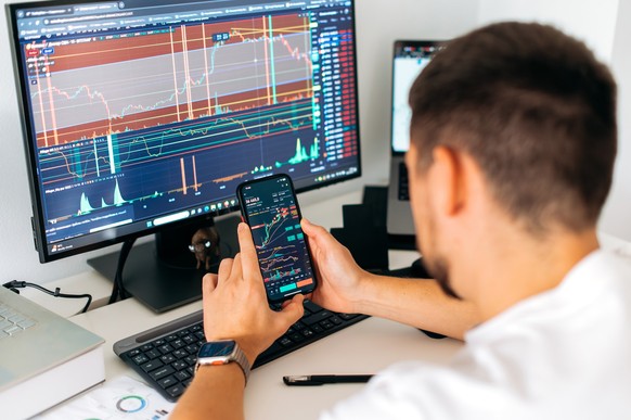 Crypto trader sits at his workplace in front of a monitor with charts. Smartphone in male hands, with trading data index chart graph on the screen, mobile phone app analytics for cryptocurrency market ...
