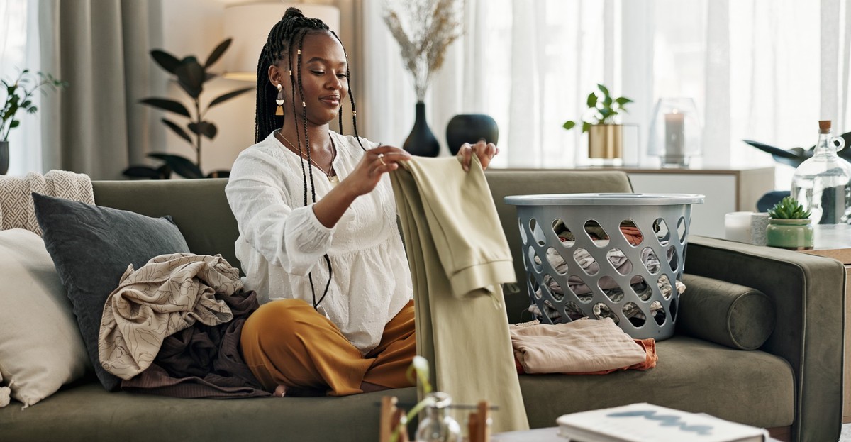 Laundry, housework and a black woman folding washing on a sofa in the living room of her home to tidy. Smile, relax and a happy young housewife cleaning her apartment for housekeeping or hygiene