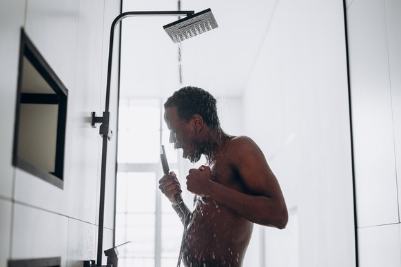Positive young African-American man sings taking shower with hot water jets in contemporary unit in bathroom close side view