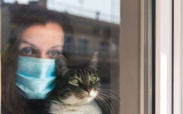 Woman with medical face mask holding cat and sadly looking out in the window holding her cat. Quarantine during Coronavirus pandemic.