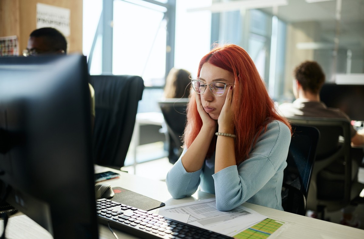 Young redhead female designer feeling exhausted while working on desktop computer in the office.