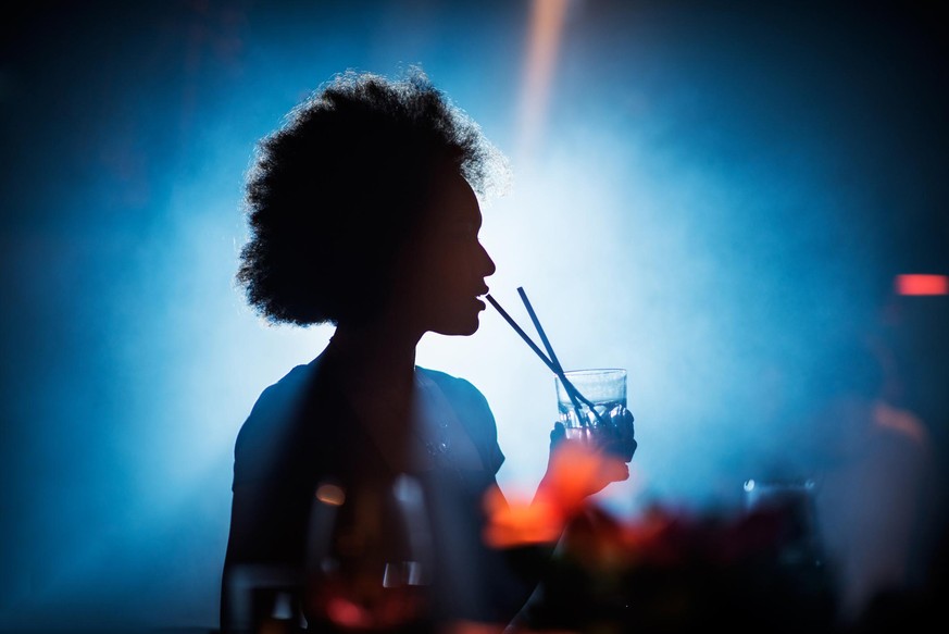 Mixed race woman having a cocktail in the bar at night out. Bright light is coming from behind.