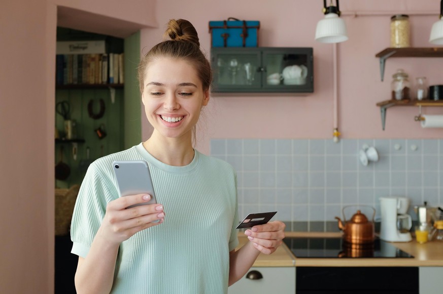 Indoor shot of young European girl pictured in her flat holding smartphone in one hand and credit card in another, processing online payments via banking application to purchase goods with content