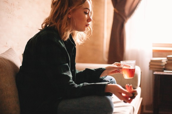 Young upset woman drinking tea and eating chocolate while sitting on sofa in living room at home