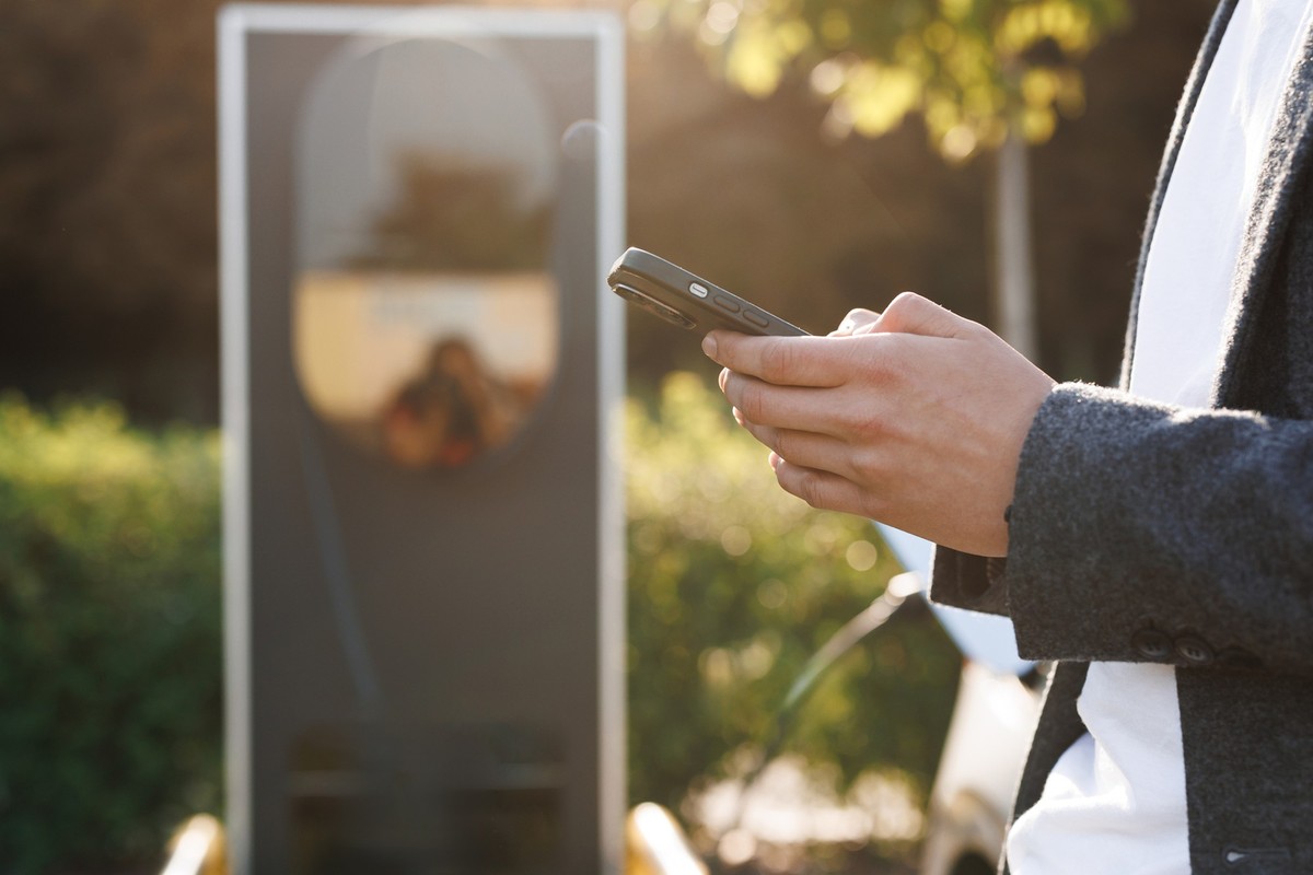 Man holding smartphone while charging car at electric vehicle charging station, closeup. Electric car charging, businessman standing in background and waiting.