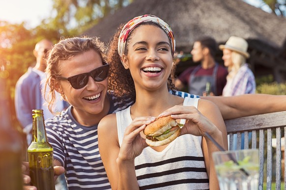 Cheerful multiethnic couple enjoying hamburger and beer at barbecue party. Happy young black woman eating burger while is sitting on boyfriend legs. Joyful guy and girl having fun at bbq party.