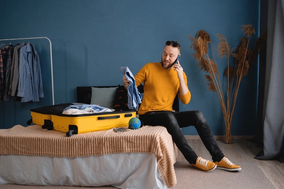 Young travel man sitting on bed near open suitcase with clothes, speaking on smartphone, booking travel tour at home, hotel reservation, buying tickets on phone. Traveler concept