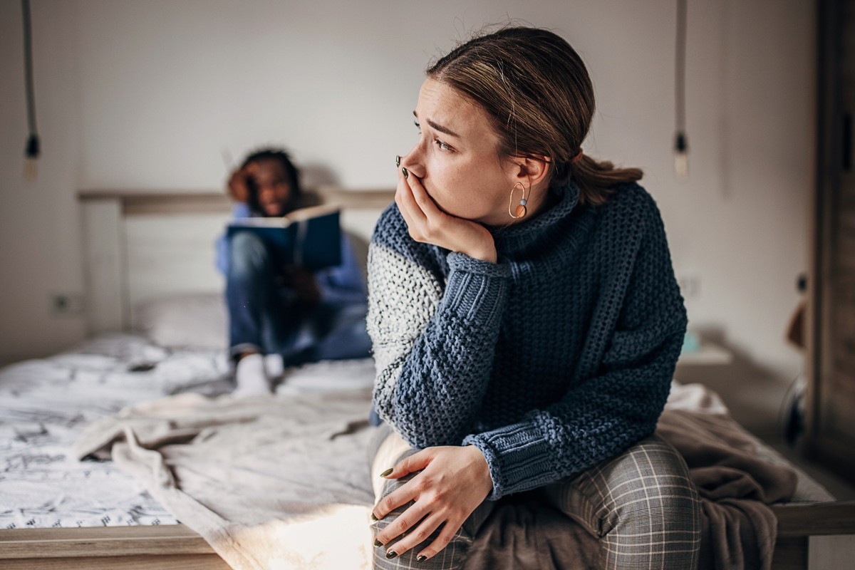 Two people, beautiful crying woman is sitting on the bed in bedroom, her boyfriend is sitting on bed behind her.