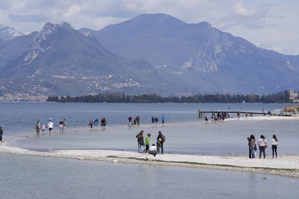 14.04.2023, Italien, Manerba del Garda: Touristen gehen über einen Streifen Land zwischen dem Festland und der Insel San Biagio, am Gardasee. Die anhaltende Trockenheit und Wasserknappheit machen Ital ...