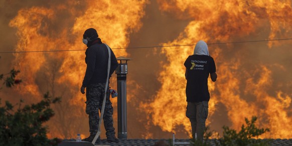 25.07.2023, Griechenland, Gennadi: Gegen Abend erreichen Flammen die Ortschaft Gennadi, Anwohner stehen auf ihren Dächern vor riesigen Flammen und versuchen mit Wasserschläuchen ihre Häuser zu retten. ...