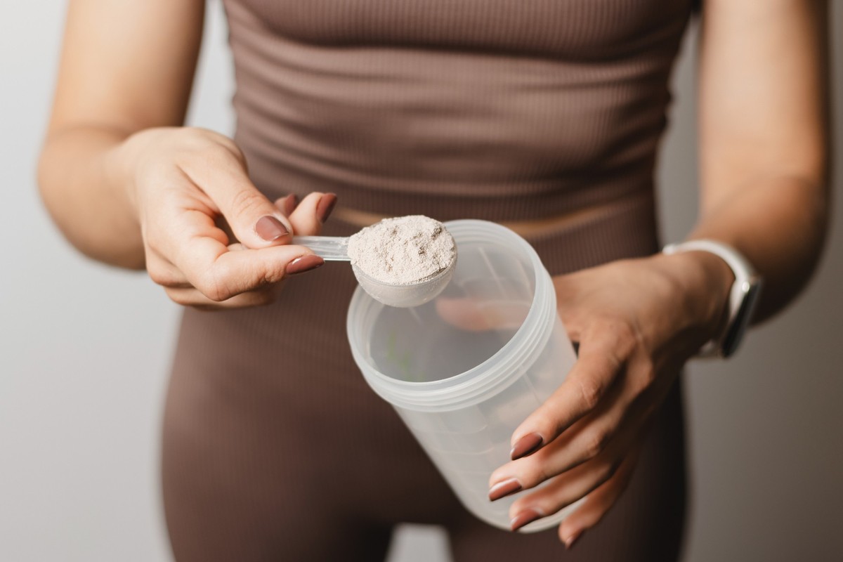 Athletic woman in sportswear with measuring spoon in her hand puts a portion of whey protein powder into a shaker, making protein drink, cropped image.