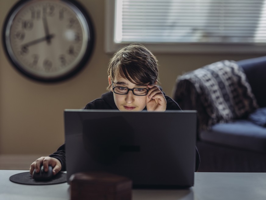 Young teenage boy with glasses looking at lap top screen. He is sitting in living room of private residence.