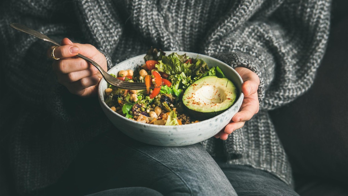 Healthy vegetarian dinner. Woman in grey jeans and sweater eating fresh salad, avocado half, grains, beans, roasted vegetables from Buddha bowl. Superfood, clean eating, dieting food concept