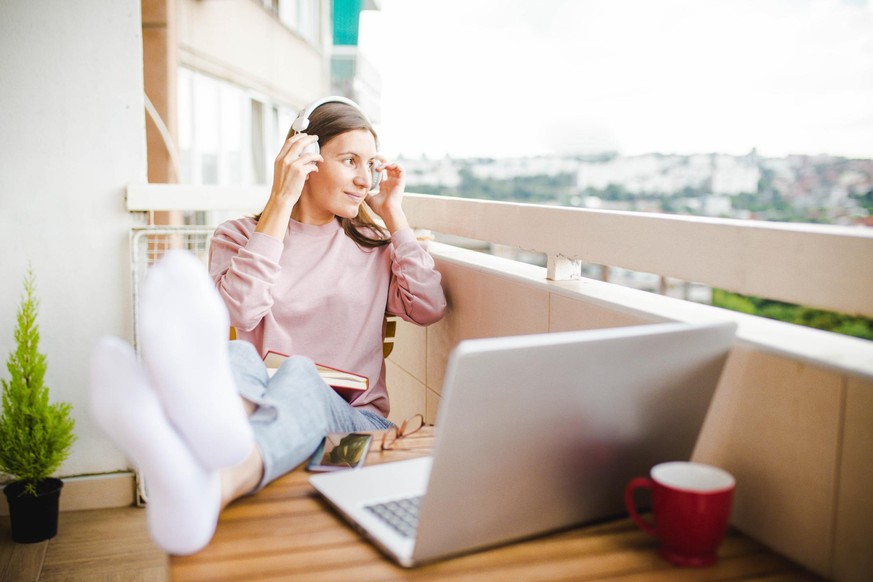 Photo of a young woman sitting on her balcony relaxing and putting wireless headphones on