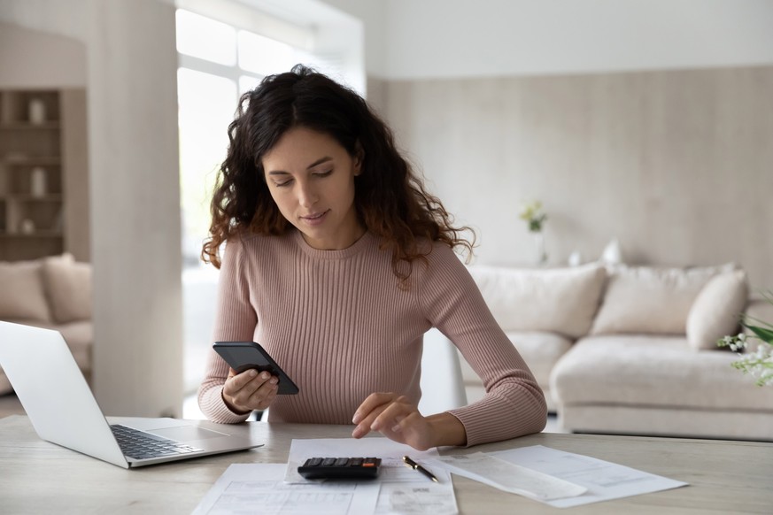 Young Hispanic woman sit at desk at home manage household finances paying bills calculating on machine cellphone. Millennial Latin female care of budget use smartphone for payment. Finance concept.