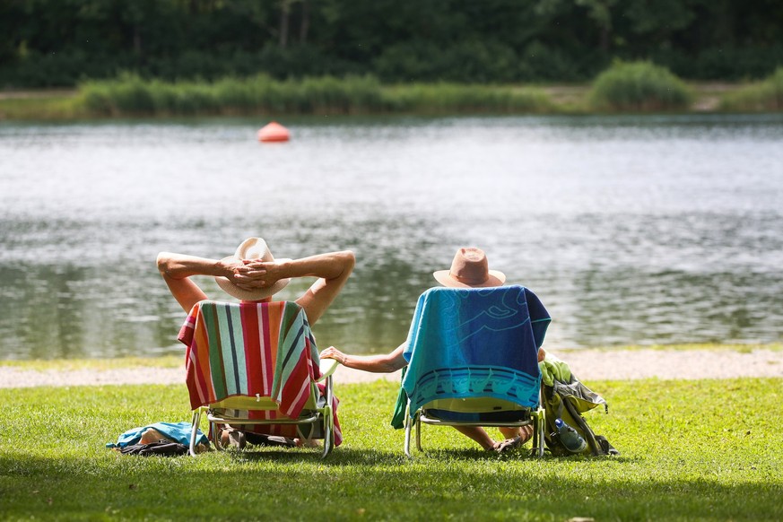 19.07.2023, Baden-Württemberg, Ertingen: Ein Mann und eine Frau mit Strohhüten sitzen an einem Badesee in ihren Stühlen in der Sonne. Bis zum Wochenende bleibt das Wetter in Baden-Württemberg sommerli ...