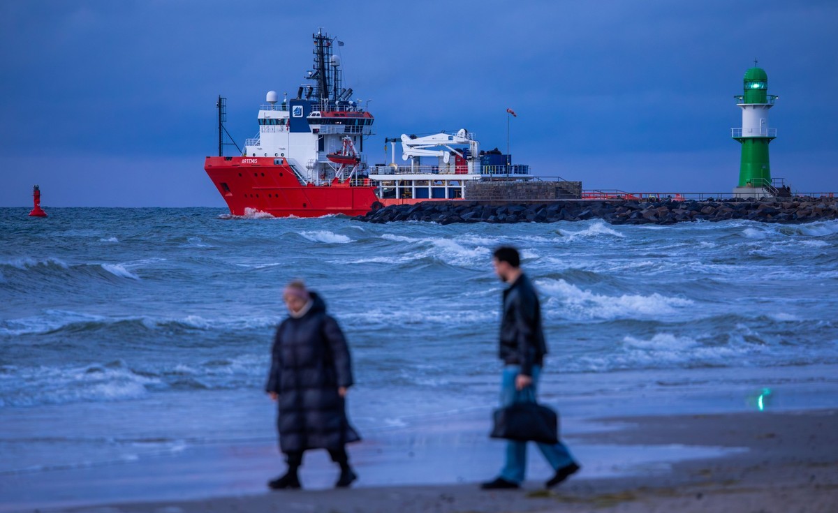 ARCHIV - 16.09.2025, Mecklenburg-Vorpommern, Rostock: Ein Frachtschiff verlässt nach Sonnenuntergang den Hafen und fährt auf die Ostsee während Spaziergänger am Strand von Warnemünde unterwegs sind. A ...