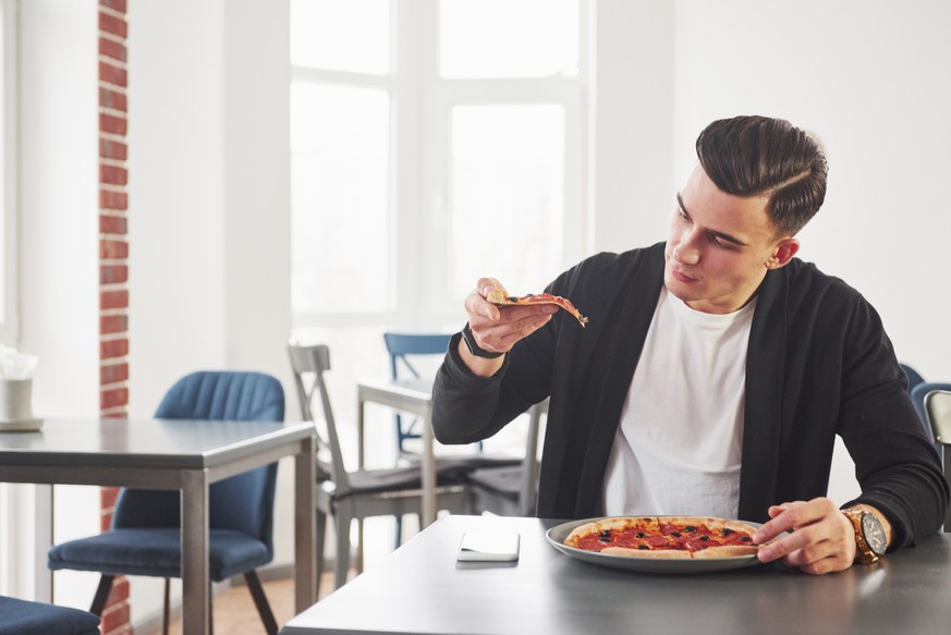 Looking at the slice. Having a breakfest with pizza at daytime. Young stylish man in black jacket.