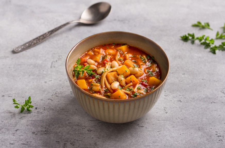 Vegan seasonal italian minestrone soup with pumpkin, beans, tomatoes, bell peppers, pasta and herbs on gray textured background. Delicious homemade food