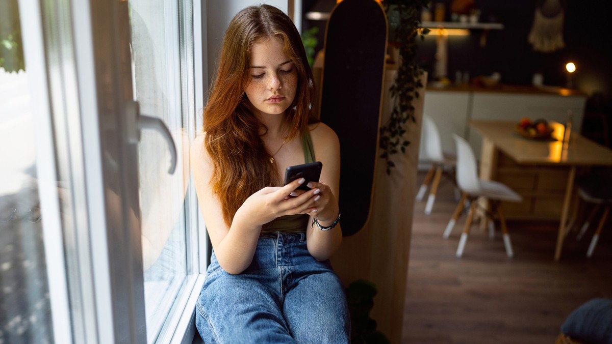 Caucasian teenage girl, sitting near a window and using mobile phone