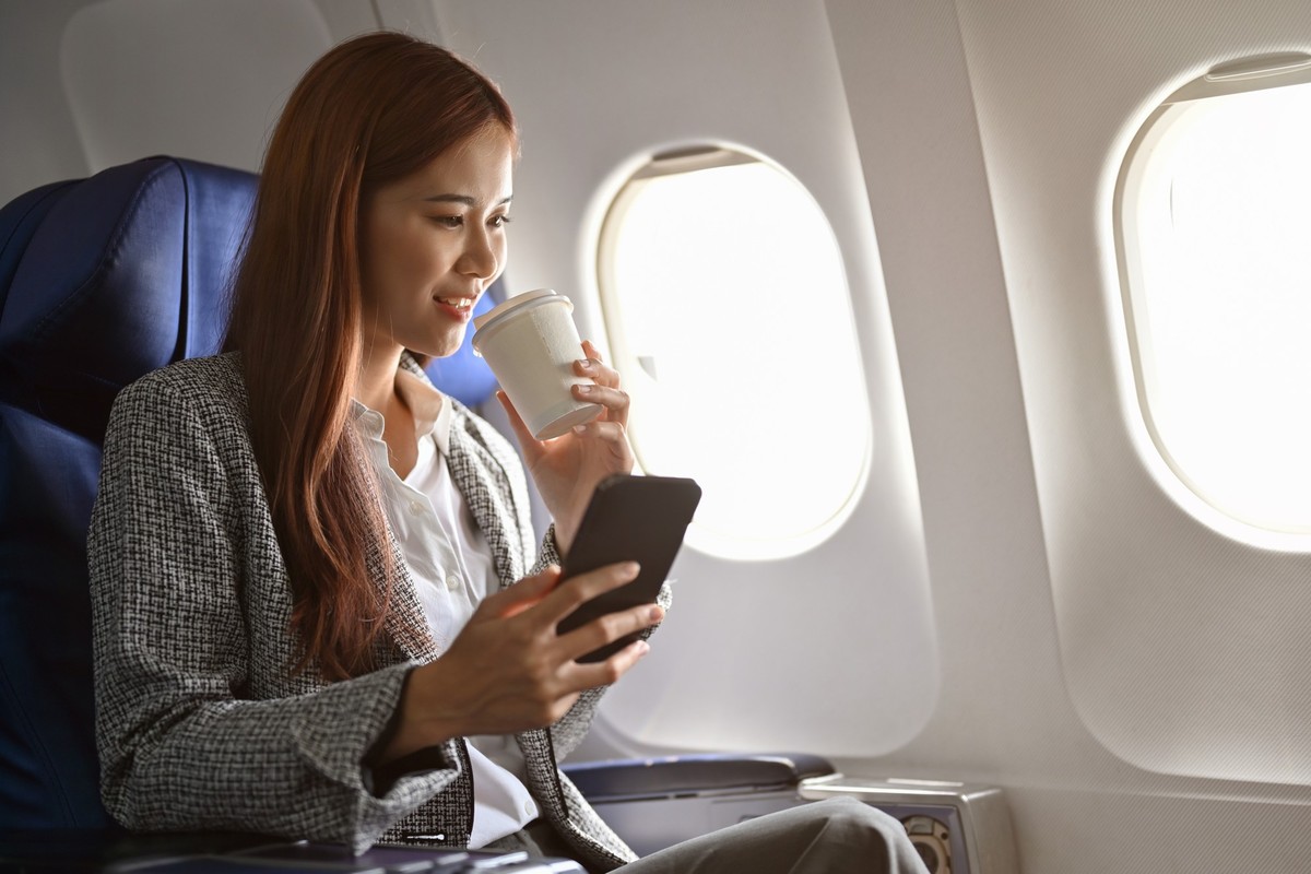 Beautiful young Asian businesswoman relaxes at her seat sipping a hot coffee and uses a modern smartphone during flight.