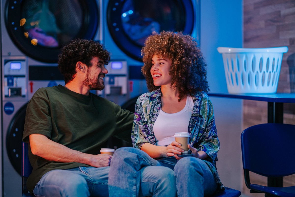 Young multi-ethnic couple drinking coffee and chatting while waiting for their laundry at a self-service laundromat