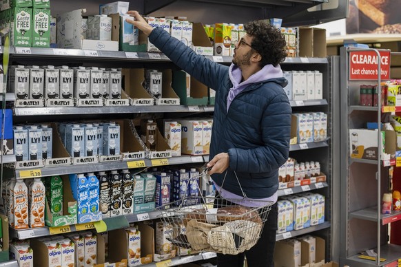 Man shopping in a supermarket while on a budget. He is looking for low prices due to inflation. He is living in the North East of England and is choosing a vegan milk.